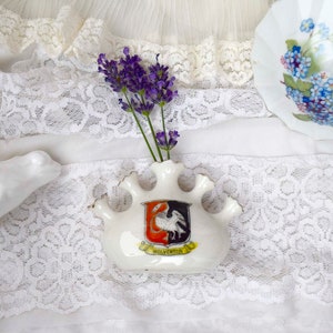 May include: A white ceramic frog ornament, a small white vase with a crest, and a decorative plate with blue flowers sit on a white lace surface. The vase holds purple lavender sprigs. The vase has the word "Wolverton" on it.