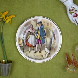 May include: A vintage ceramic plate with a colorful illustration of a couple walking a dog in a city setting. The plate has a gold rim and the text "Our of London, 18th Century" on the bottom.