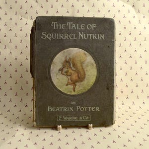 Puede incluir: Un libro antiguo de tapa dura titulado "The Tale of Squirrel Nutkin" de Beatrix Potter. La portada presenta una ardilla marrón con una cola tupida que sostiene una nuez en sus patas. El libro está publicado por F. Warne & Co.