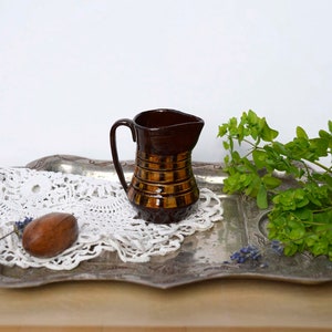 May include: A small, brown ceramic jug with a handle and horizontal gold bands. The jug sits on a silver tray with a white lace doily, a wooden object, and green foliage. The background is white.