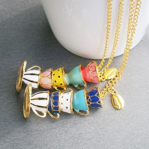 Puede incluir: Un collar de cadena dorada con un dije de tazas de té apiladas en varios colores, incluyendo rosa, amarillo, azul y blanco. El dije está unido a un dije de grano de café dorado.
