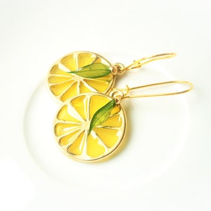 Puede incluir: Un par de pendientes de oro con un diseño de rodaja de limón. Cada pendiente presenta una rodaja de limón amarilla con una hoja verde en su interior. Los pendientes están sobre un fondo blanco.