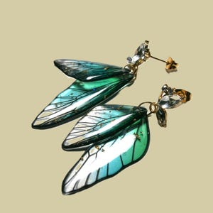 May include: A pair of gold-coloured stud earrings with iridescent, wing-shaped pendants in teal, green, and white. Each earring has a cluster of clear, faceted crystals. The earrings are designed to resemble fairy wings.