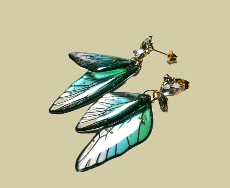 May include: A pair of gold-coloured stud earrings with iridescent, wing-shaped pendants in teal, green, and white. Each earring has a cluster of clear, faceted crystals. The earrings are designed to resemble fairy wings.