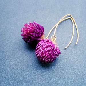 May include: A pair of dangle earrings featuring vibrant purple flower-shaped beads. The earrings have gold-colored, curved ear wires. The background is a solid, muted blue.