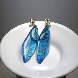 May include: A pair of blue and gold-flecked earrings shaped like butterfly wings. The earrings have a gold-coloured top with a small, clear crystal butterfly accent. The earrings are displayed on a white, round dish.