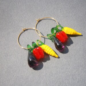 May include: A pair of gold hoop earrings with dangling charms shaped like vegetables. The charms are a red pepper, a yellow pepper, a green pepper, a purple eggplant, and a yellow corn cob.