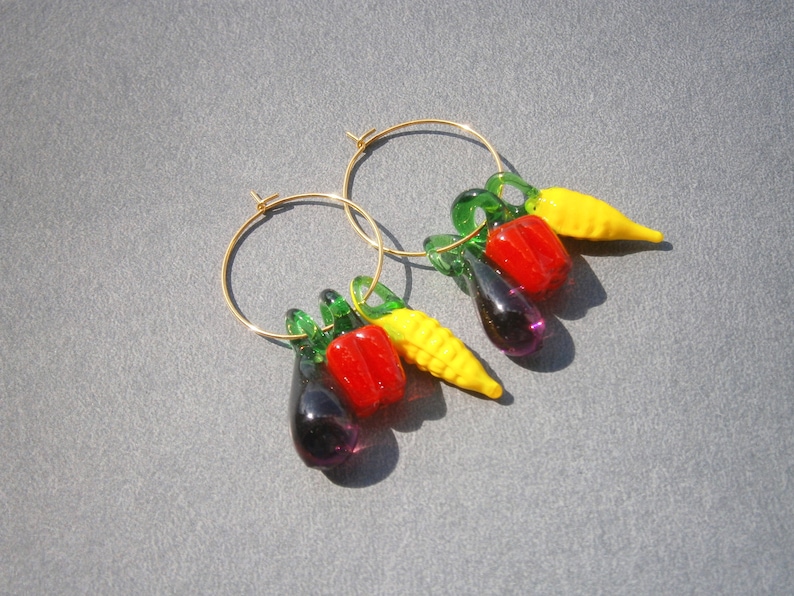 May include: A pair of gold hoop earrings with dangling charms shaped like vegetables. The charms are a red pepper, a yellow pepper, a green pepper, a purple eggplant, and a yellow corn cob.