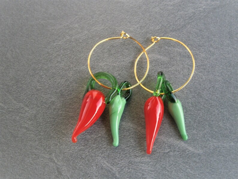 May include: A pair of gold hoop earrings with red and green glass chili pepper charms. The charms are detailed with green stems and leaves.