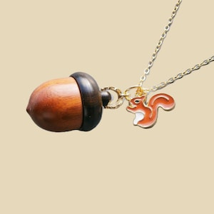 Op de afbeelding: Een goudkleurige ketting met een hanger met een houten eikel en een kleine, oranje en witte eekhoornbedel. De eikel is bruin met een donkerbruine dop. De eekhoornbedel is gedetailleerd met witte accenten.