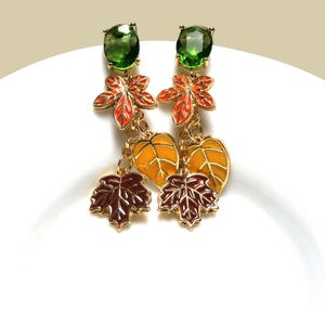 May include: A pair of gold-coloured earrings with a fall theme. Each earring features a green gemstone, orange and red leaf, yellow leaf, and a brown leaf. The earrings are designed with a dangle style.