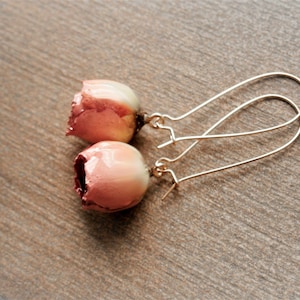 Könnte beinhalten: Ein Paar Ohrhänger mit zwei kleinen, abgerundeten, rosafarbenen Blütenknospen mit glatter, glänzender Oberfläche. Jede Knospe ist an einem dünnen, geschwungenen, goldfarbenen Metalldraht-Ohrhaken befestigt, der nach oben verläuft.