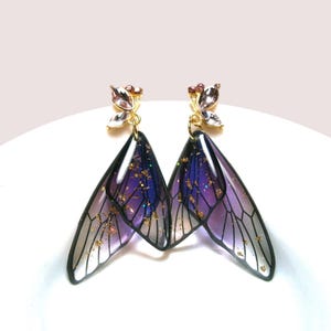 May include: A pair of handcrafted earrings with a wing design. The wings have a purple and blue gradient with gold flecks, black outlines, and gold and crystal accents. The earrings are presented on a white surface.