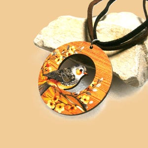 May include: Wooden pendant necklace with a bird design. The circular pendant has a bird on a branch with yellow flowers. The necklace has a brown and black cord. The bird has a white face and orange accents. The background is a light tan colour.