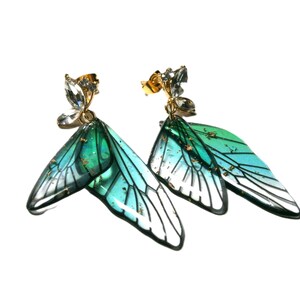May include: A pair of gold-coloured stud earrings featuring a butterfly wing design. The wings are a gradient of teal and green with black outlines and gold flecks. The studs are decorated with clear crystals.