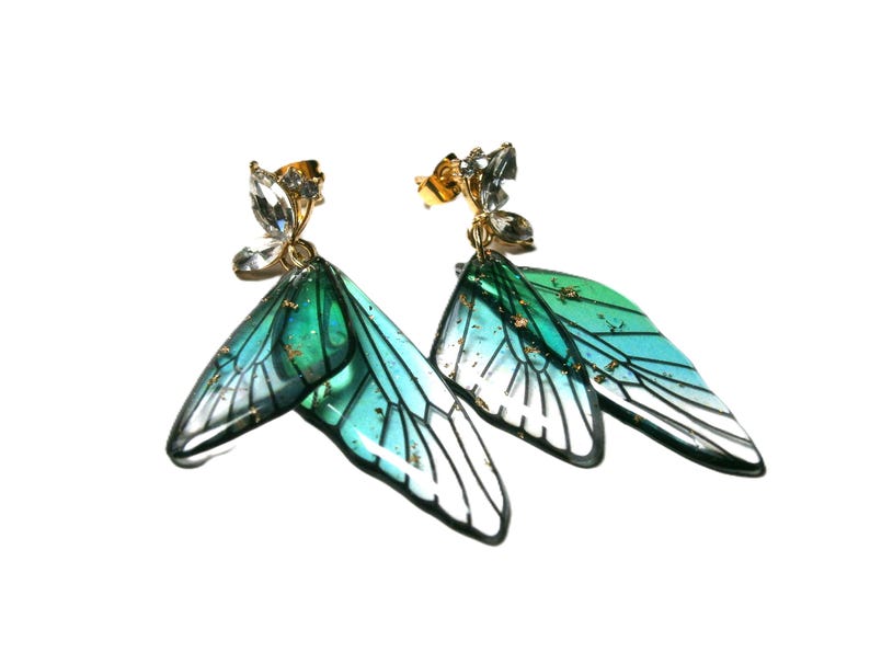 May include: A pair of gold-coloured stud earrings featuring a butterfly wing design. The wings are a gradient of teal and green with black outlines and gold flecks. The studs are decorated with clear crystals.
