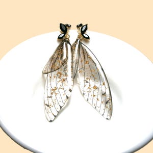 May include: A pair of transparent wing-shaped earrings. The earrings have black accents and gold flecks throughout the wing design. The wings are detailed with black lines, creating a unique and ethereal aesthetic. The earrings are on a white surface.