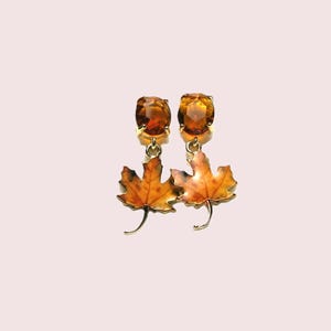 May include: A pair of drop earrings featuring faceted amber-coloured stones set in gold-toned metal. Each earring suspends a detailed maple leaf charm with a gradient of orange, yellow, and brown tones, mimicking the appearance of autumn foliage.