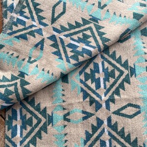 Pu&ograve; includere: Una coperta piegata con un motivo geometrico del sud-ovest in verde acqua, bianco e verde acqua scuro su sfondo beige. La coperta ha un aspetto morbido e strutturato ed &egrave; piegata per mostrare il motivo.