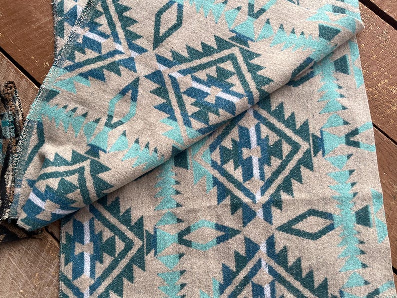 Pu&ograve; includere: Una coperta piegata con un motivo geometrico del sud-ovest in verde acqua, bianco e verde acqua scuro su sfondo beige. La coperta ha un aspetto morbido e strutturato ed &egrave; piegata per mostrare il motivo.