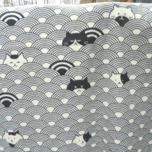May include: A rectangular blanket featuring a repeating pattern of stylized waves in shades of gray and white. The design incorporates playful illustrations of cats peeking out from behind the waves, creating a whimsical and cozy aesthetic.