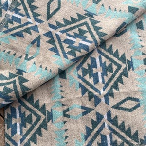 Pu&ograve; includere: Una coperta beige piegata con un motivo geometrico in verde acqua e verde acqua scuro. La coperta ha un aspetto morbido e strutturato e un bordo sfrangiato. Il design presenta forme ripetute di diamanti e frecce.