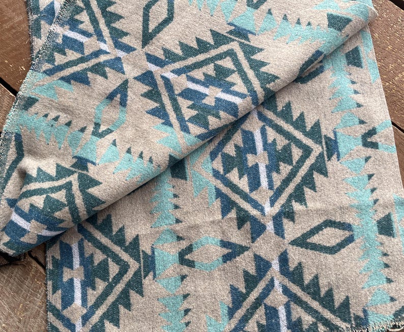 Pu&ograve; includere: Una coperta beige piegata con un motivo geometrico in verde acqua e verde acqua scuro. La coperta ha un aspetto morbido e strutturato e un bordo sfrangiato. Il design presenta forme ripetute di diamanti e frecce.