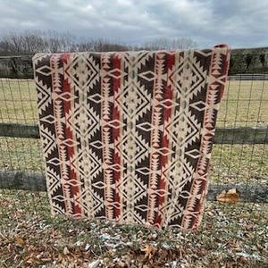 May include: A Southwestern-style blanket with a geometric pattern in shades of brown, cream, and red. The blanket is draped over a wooden fence, showcasing its intricate design and textured weave. The blanket's dimensions are not visible.