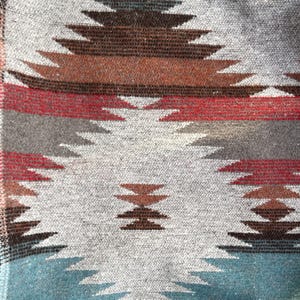 May include: Close-up of a woven textile with a geometric pattern. The design features repeating diamond shapes in shades of brown, red, gray, and blue on a light gray background. The textile has a textured appearance.