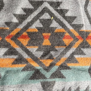 May include: Close-up of a woven textile with a geometric pattern. The design features gray, orange, red, and teal triangles and diamond shapes on a gray background. The textile has a textured appearance.