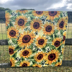May include: A yellow blanket with a repeating pattern of sunflowers. The sunflowers have yellow petals and brown centers, with green leaves and stems. The blanket is draped over a wooden fence.