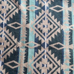 May include: Close-up of a textile with a geometric pattern in shades of teal, navy, and tan. The design features diamond and zigzag shapes, creating a Southwestern or tribal aesthetic. The fabric appears to be woven.