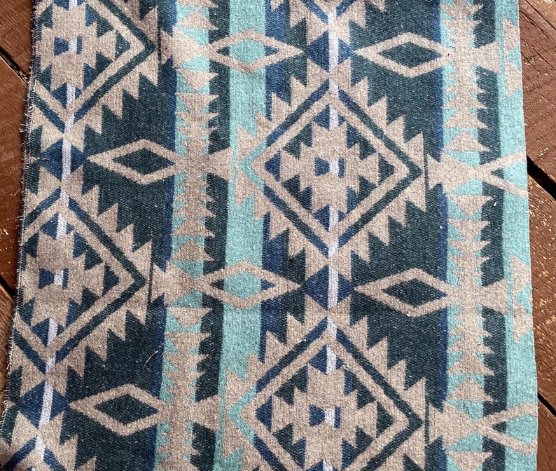 Pu&ograve; includere: Primo piano di un tessuto con un motivo geometrico nei toni del verde acqua, blu navy e beige. Il design presenta forme a diamante e a zig-zag, creando un'estetica sud-ovest o tribale. Il tessuto sembra essere tessuto.