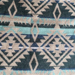 Pu&ograve; includere: Primo piano di un tessuto con un motivo geometrico nei toni del beige, verde acqua, blu navy e bianco. Il design presenta forme a diamante e triangolo, creando un'estetica sud-occidentale o tribale. Il tessuto sembra essere tessuto.