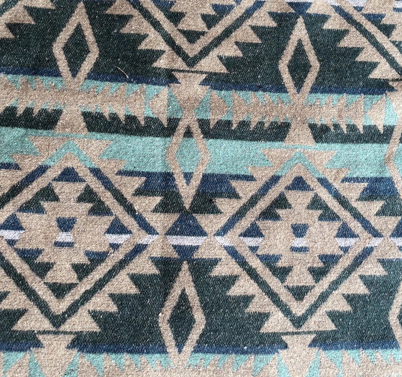 Pu&ograve; includere: Primo piano di un tessuto con un motivo geometrico nei toni del beige, verde acqua, blu navy e bianco. Il design presenta forme a diamante e triangolo, creando un'estetica sud-occidentale o tribale. Il tessuto sembra essere tessuto.