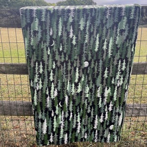 May include: A rectangular fabric item with a forest pattern of green and white trees on a black background. The design includes crescent moons and full moons. The item is draped over a fence.