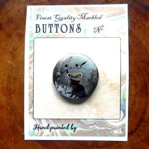 May include: A marbled button with a black and gray design featuring a silhouette of a cat and birds. The card reads "Finest Quality Marbled Buttons" and "Hand painted by".