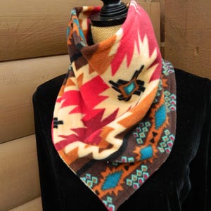 May include: A colorful fleece neck warmer with a geometric pattern in shades of red, orange, brown, turquoise, and white. The neck warmer is folded and displayed on a mannequin.