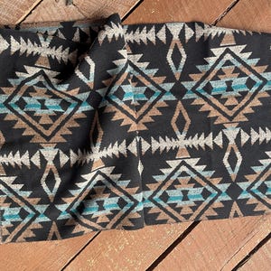 May include: A folded black fabric with a geometric pattern in shades of brown, teal, and off-white. The design features diamond and arrow shapes. The fabric is likely a scarf or wrap.