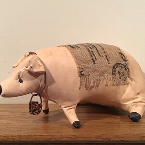 May include: A pink pig figurine with a brown burlap sack on its back. The pig has a small brown basket hanging from its neck and a pink curly tail.