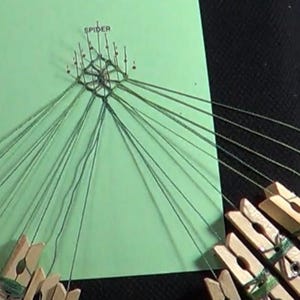 Puede incluir: Un patrón de telaraña de cuerda verde se está creando en una hoja de papel verde con la palabra "SPIDER" escrita en la parte superior. Las cuerdas están sujetas a pinzas para la ropa.