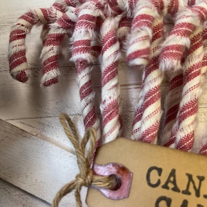 May include: Close-up of several fabric candy canes with red and white stripes. The candy canes are tied together with twine and a tag that says "CAN". The fabric has a frayed texture, and the background is a light-colored wooden surface.