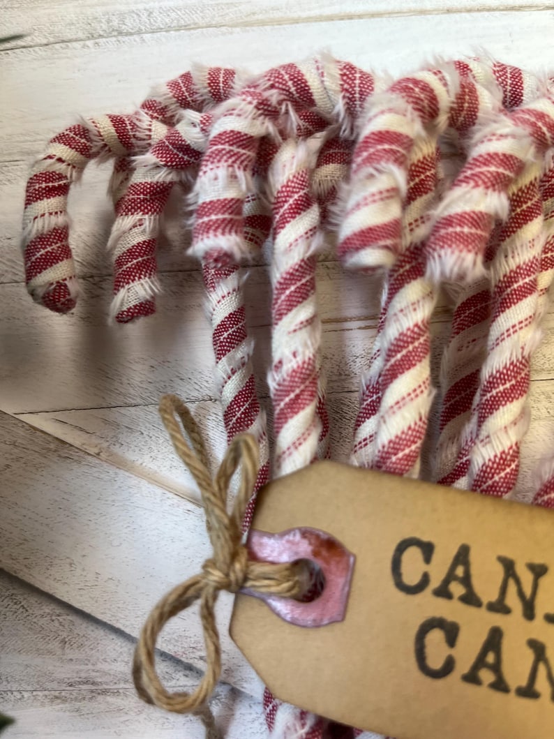 May include: Close-up of several fabric candy canes with red and white stripes. The candy canes are tied together with twine and a tag that says "CAN". The fabric has a frayed texture, and the background is a light-colored wooden surface.