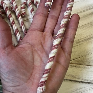 May include: A red and white striped fabric candy cane. The candy cane is held in a hand, with several others visible in the background. The fabric has a twisted design, and the ends are frayed.