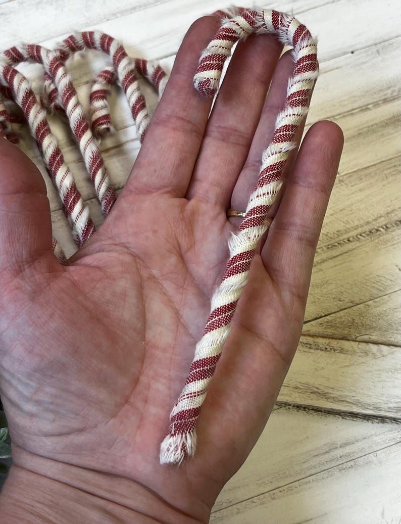 May include: A red and white striped fabric candy cane. The candy cane is held in a hand, with several others visible in the background. The fabric has a twisted design, and the ends are frayed.