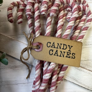 May include: A bundle of fabric candy canes with red and white stripes. A brown tag tied with twine reads "CANDY CANES" in black letters. The candy canes are arranged in a curved shape, suggesting a festive holiday theme.