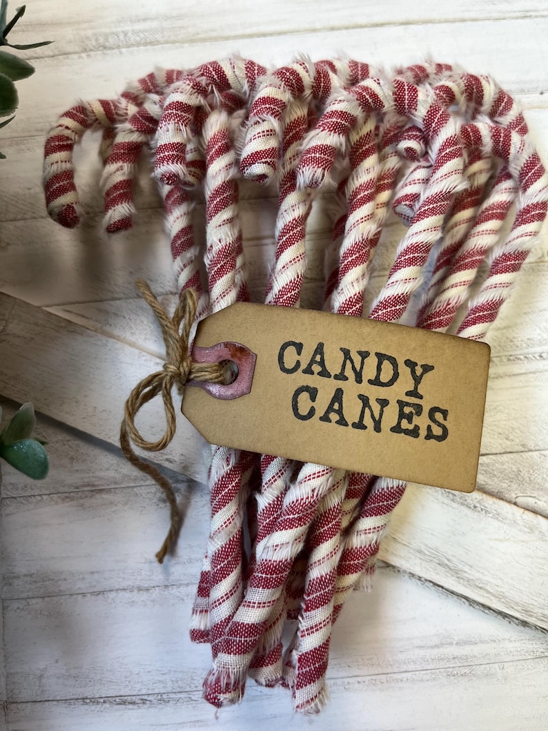 May include: A bundle of fabric candy canes with red and white stripes. A brown tag tied with twine reads "CANDY CANES" in black letters. The candy canes are arranged in a curved shape, suggesting a festive holiday theme.