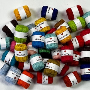 May include: A collection of colorful cotton yarn skeins. Each skein is wrapped with a white label that reads "Cotton Yarn" with a heart symbol. The yarn colors include blue, red, yellow, green, and brown.