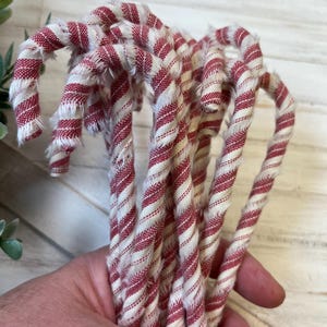 May include: A bundle of fabric candy canes with red and white stripes. The fabric has a frayed edge, giving a rustic appearance. The candy canes are curved and appear to be handmade, perfect for holiday decor.