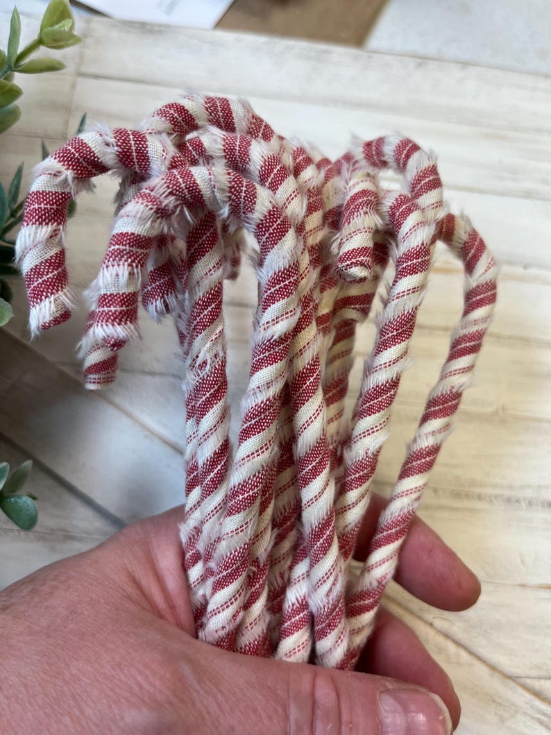May include: A bundle of fabric candy canes with red and white stripes. The fabric has a frayed edge, giving a rustic appearance. The candy canes are curved and appear to be handmade, perfect for holiday decor.